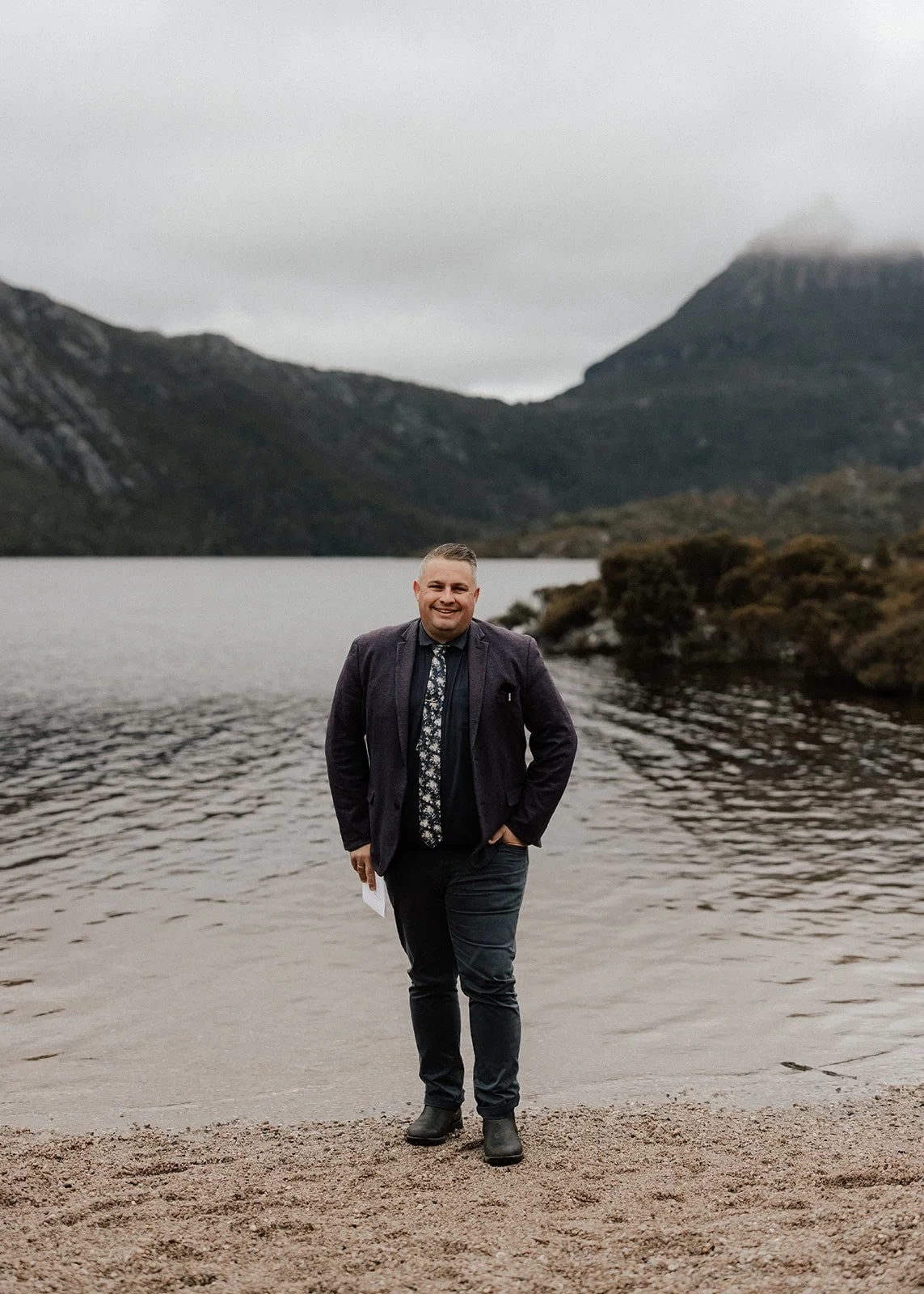 Celebrant Josh Withers standing on the lakeshore beneath misty mountain peaks, at Dove Lake, Cradle Mountain, Tasmania, for an Elopement Collective elopement. Photo by Bulb Creative.