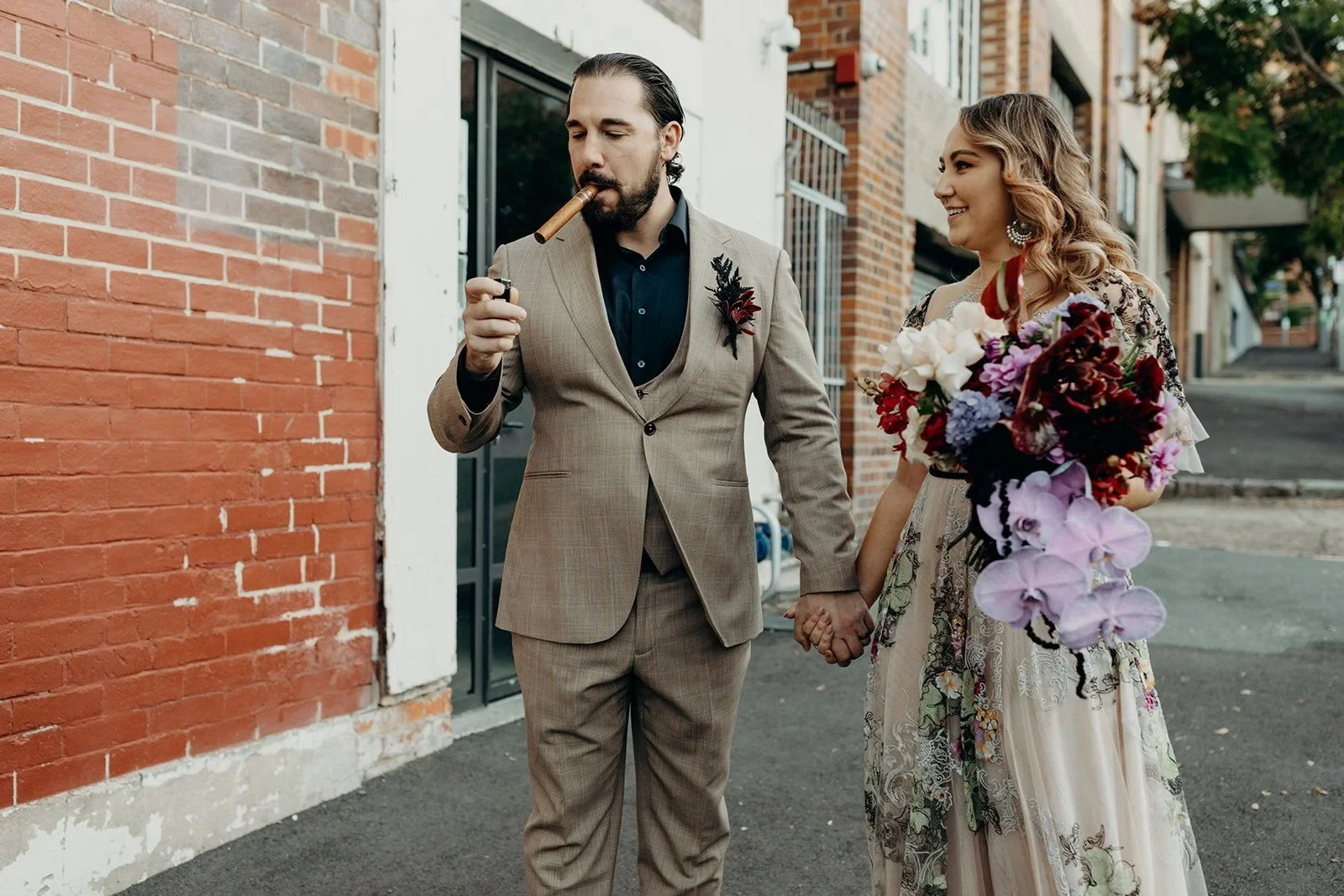 Daniel smoking a cigar as Victoria smiles beside him with her bouquet by Mrs Gibbons Flowers on a brick-lined street, in Fortitude Valley, Brisbane, for an Elopement Collective elopement. Photo by Tessa Shannon.