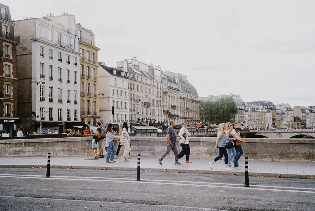 Street scene in Paris with people walking near historic buildings and a bridge over the Seine River.