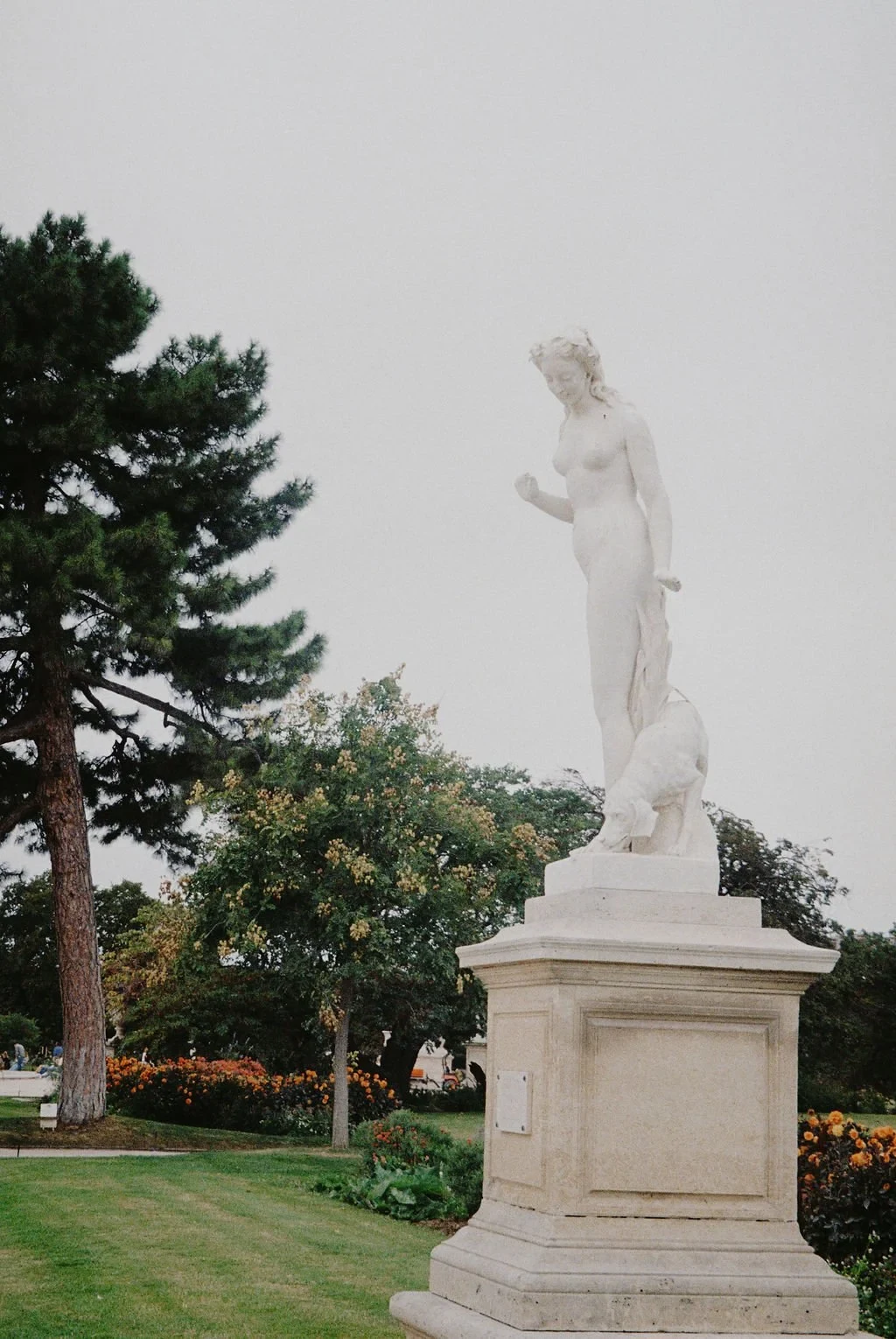 Classic marble sculpture of a nude woman on pedestal in a garden with trees and flowers.