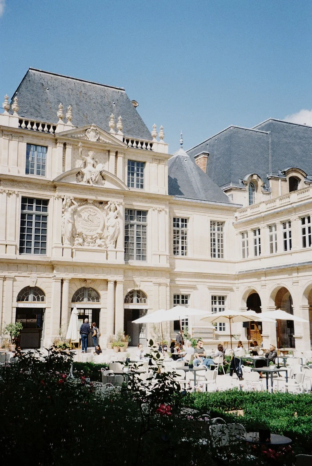 Historic building with detailed stone facade, people sitting at outdoor café tables, blue sky background.
