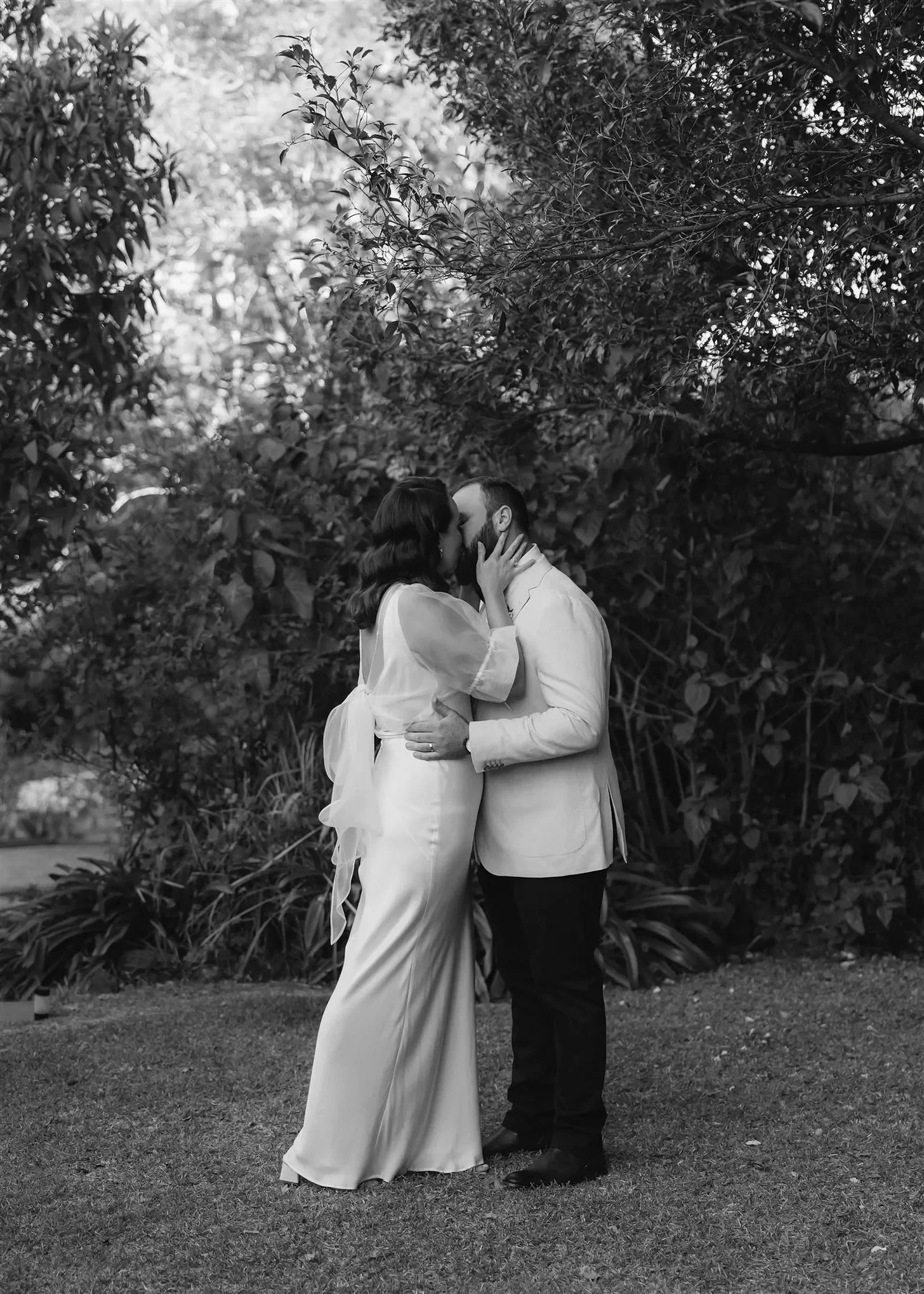 Black-and-white photograph of Tineeka and Chris kissing on the garden lawn, in Montville, Sunshine Coast Hinterland, Queensland, for an Elopement Collective elopement. Photo by Bulb Creative.