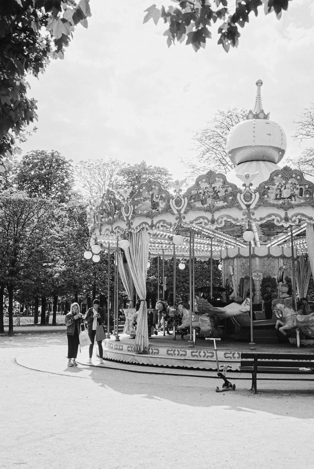 Black and white photo of a vintage carousel in a park with people standing nearby and trees in the background.