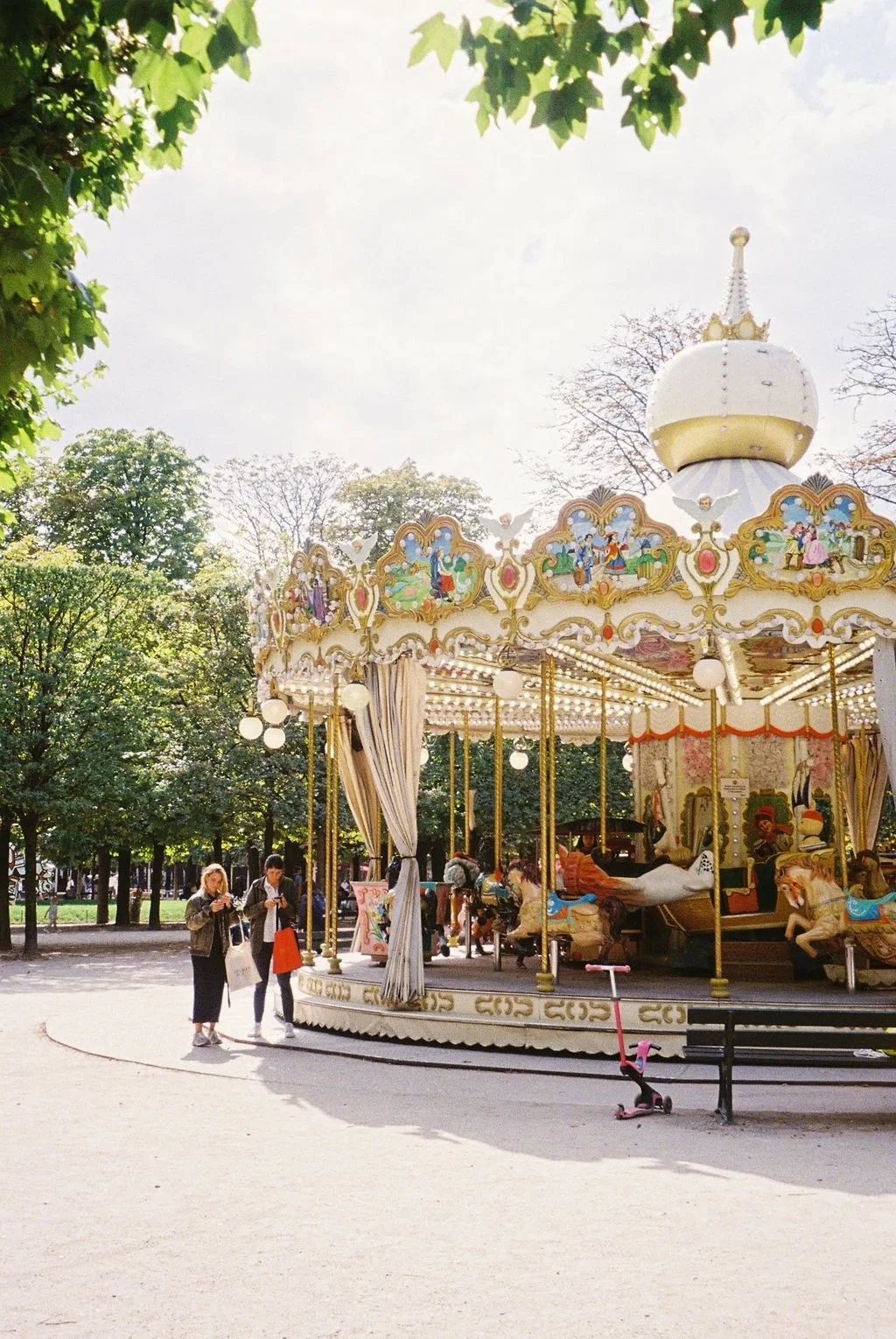 Vintage carousel in a park surrounded by trees, with people standing nearby and a child's scooter on the ground.