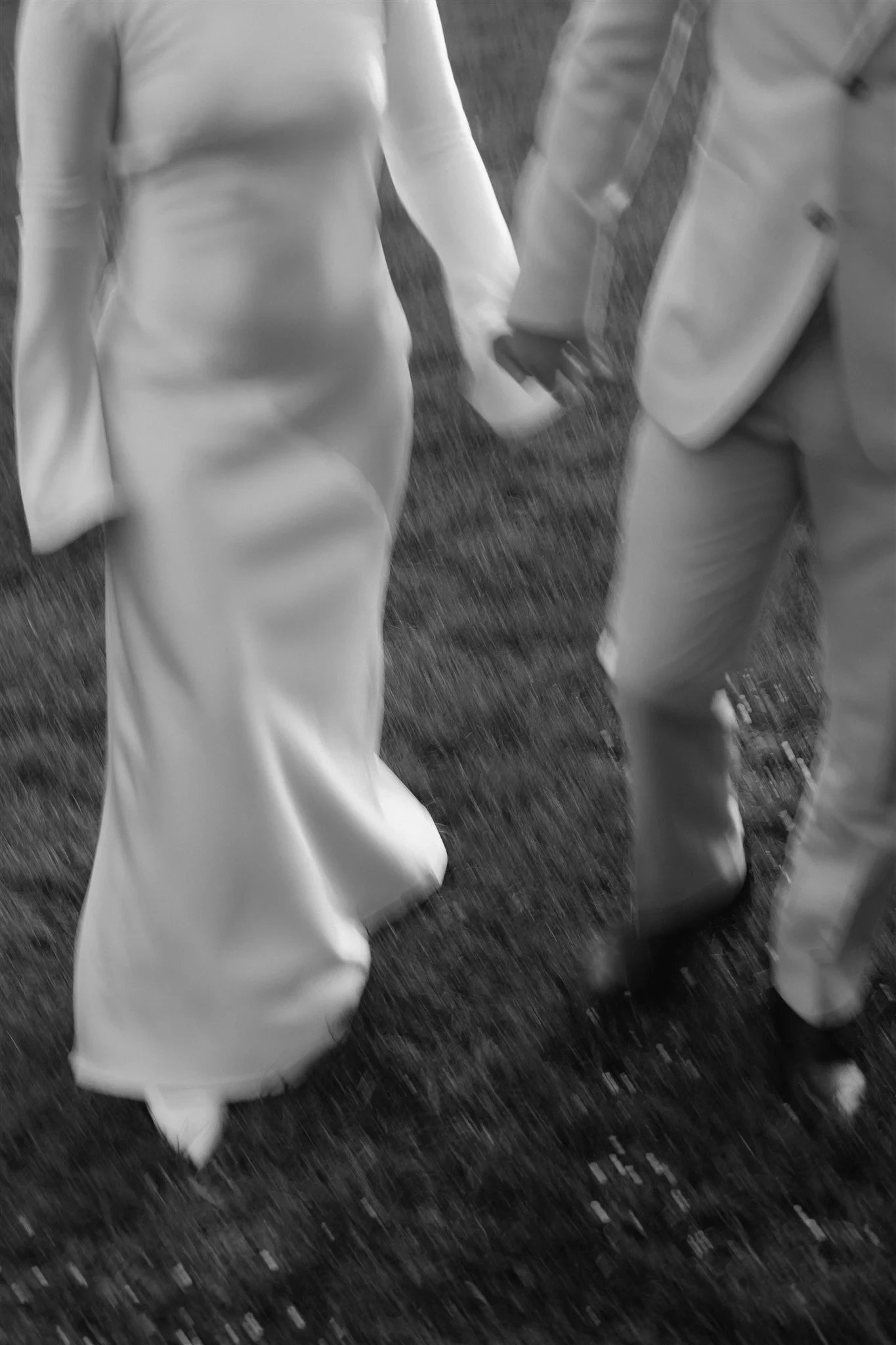 Black-and-white detail of Maddy’s dress and Cameron’s suit as they walk across the grass, at The Mayor’s Residence in Daylesford, Victoria, for an Elopement Collective elopement. Photo by Bulb Creative.