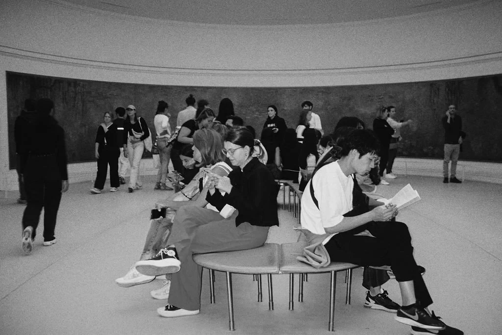 Black and white photo of people in an art gallery, some sitting and reading or using phones, with a large painting on the wall.