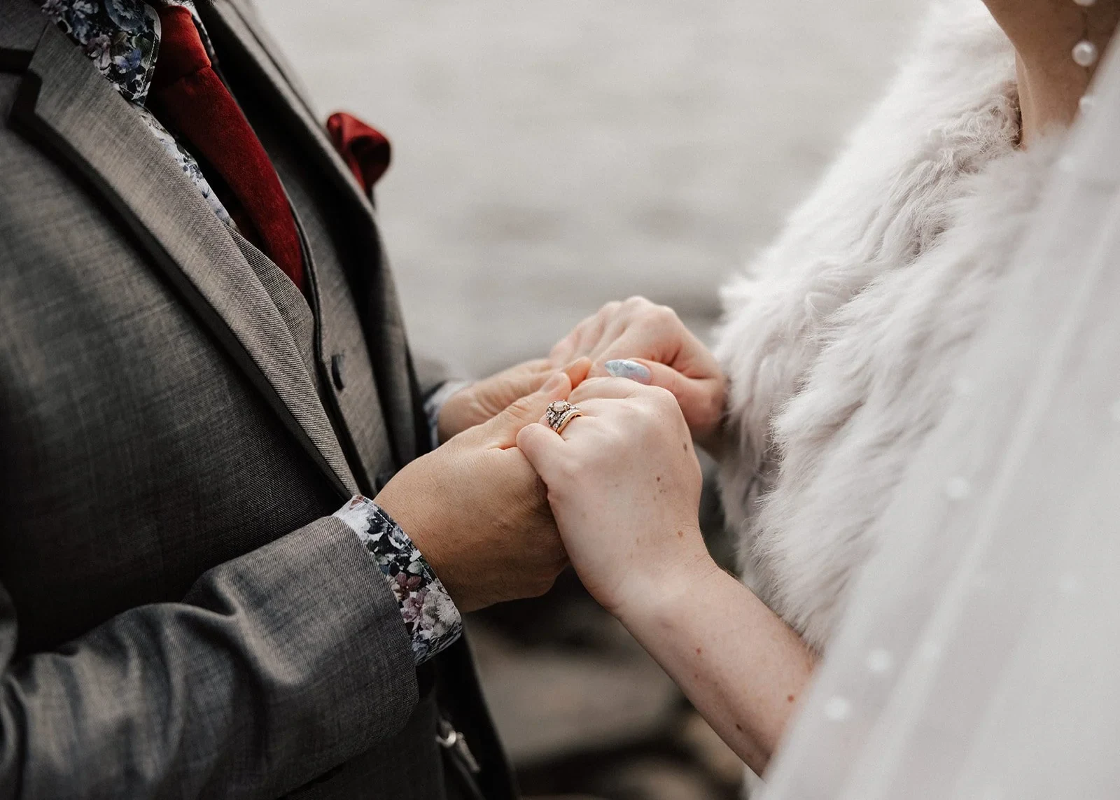 Close-up of Sarah and Jono holding hands, with their wedding rings, his grey suit and her textured wrap visible, at Pumphouse Point, Lake St Clair, Tasmania, for an Elopement Collective elopement. Photo by Bulb Creative.
