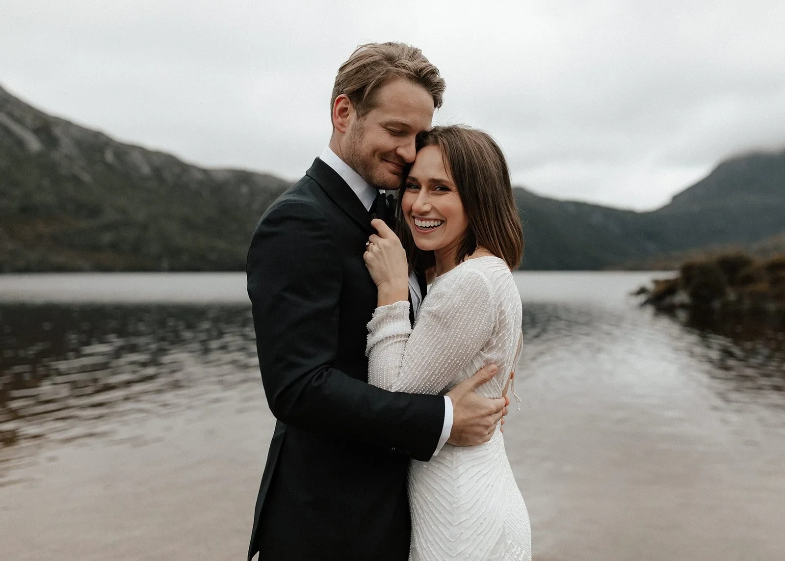 April laughing against Josh’s chest as he holds her close on the lakeshore, at Dove Lake, Cradle Mountain, Tasmania, for an Elopement Collective elopement. Photo by Bulb Creative.