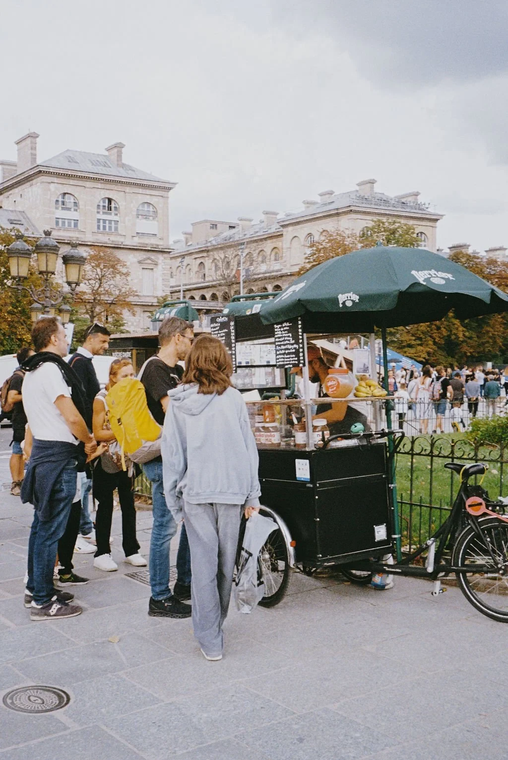 People standing in line at a street food cart with a green umbrella in an urban area, historical buildings in the background.