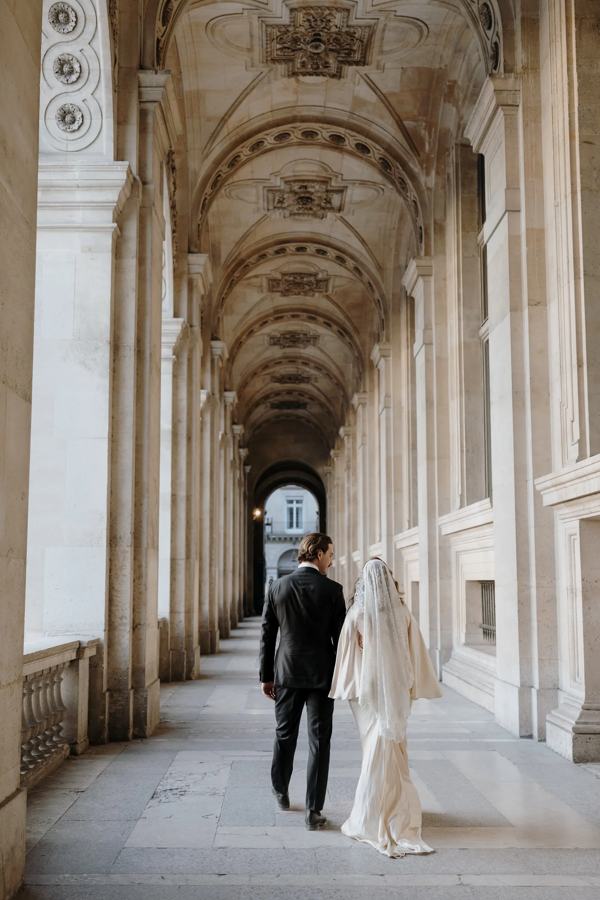 Couple in wedding attire walking through an ornate stone arcade