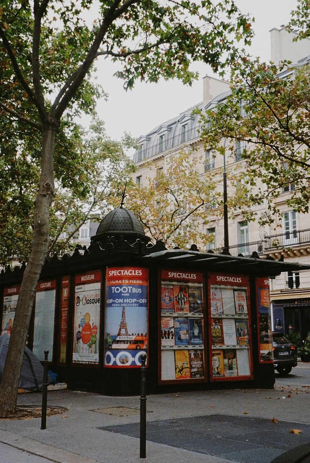 Kiosk in Paris surrounded by trees and historical buildings, featuring posters for cultural events and tours.