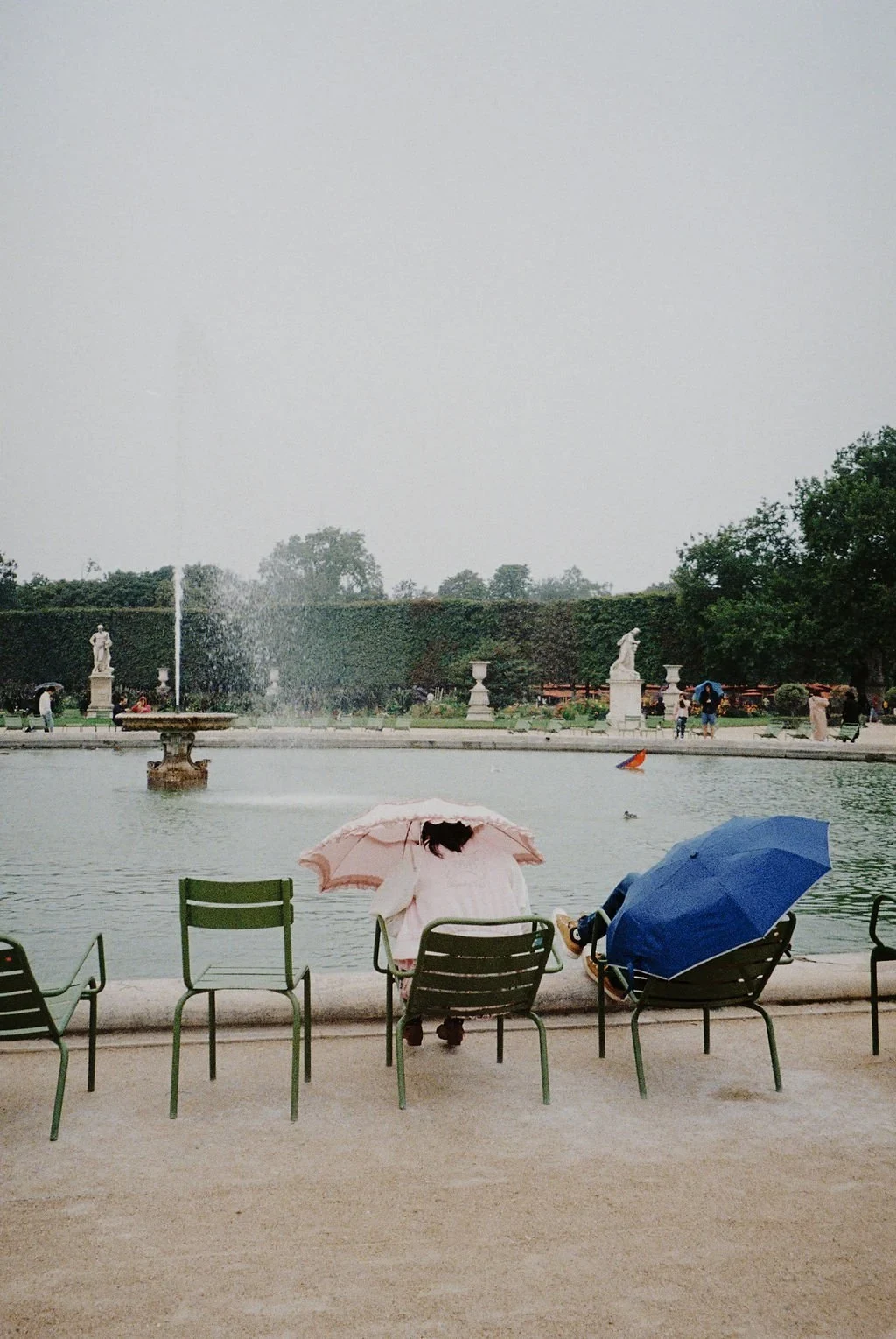 People sitting on green chairs by a fountain in a garden, one holding a pink umbrella and another with a blue one, statues and trees in the background.
