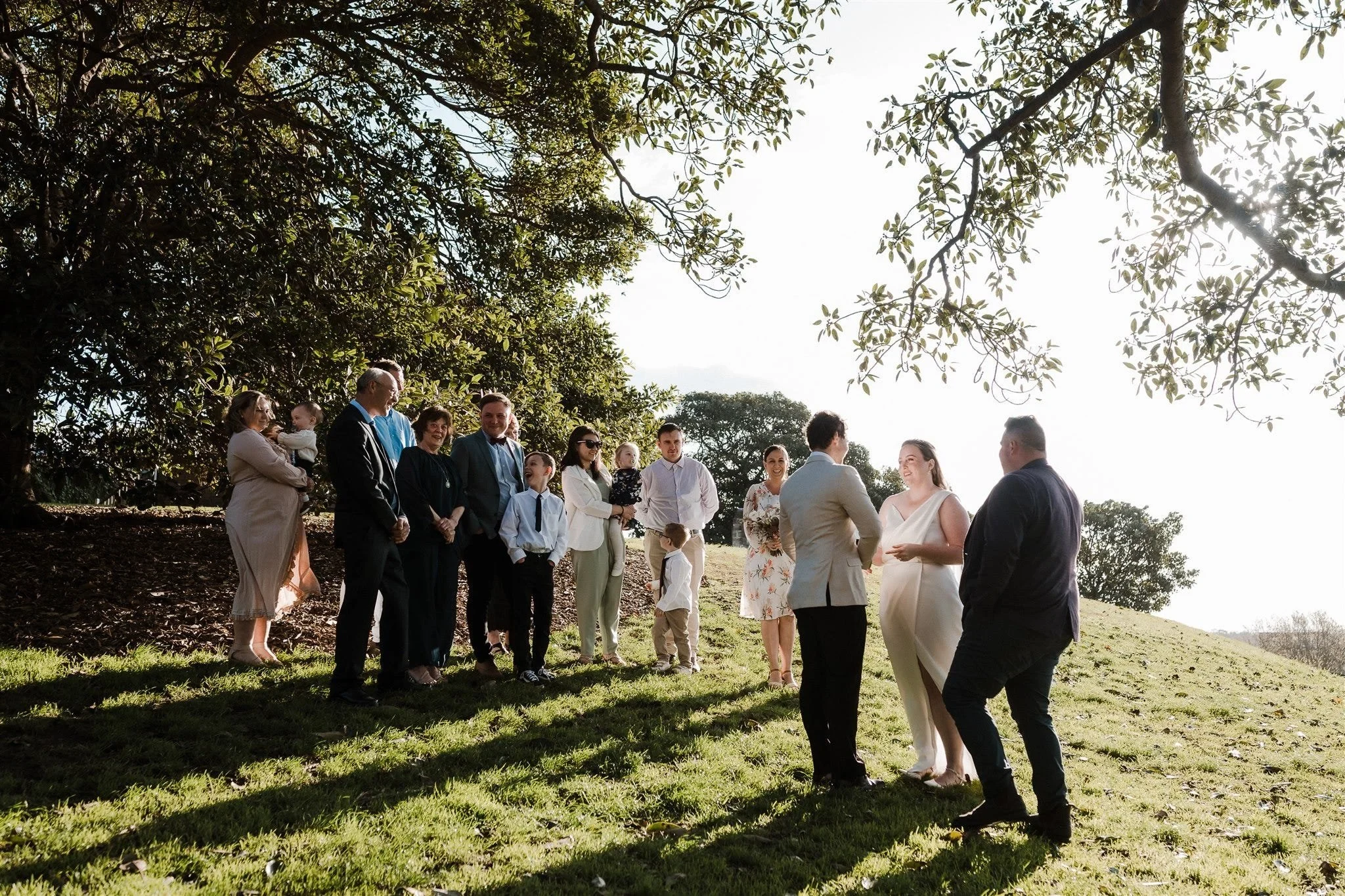 Alyce and Andrew exchanging vows with celebrant Josh Withers and guests gathered beneath a large tree, at Observatory Hill in Sydney, New South Wales, for an Elopement Collective elopement. Photo by Jason Corroto.